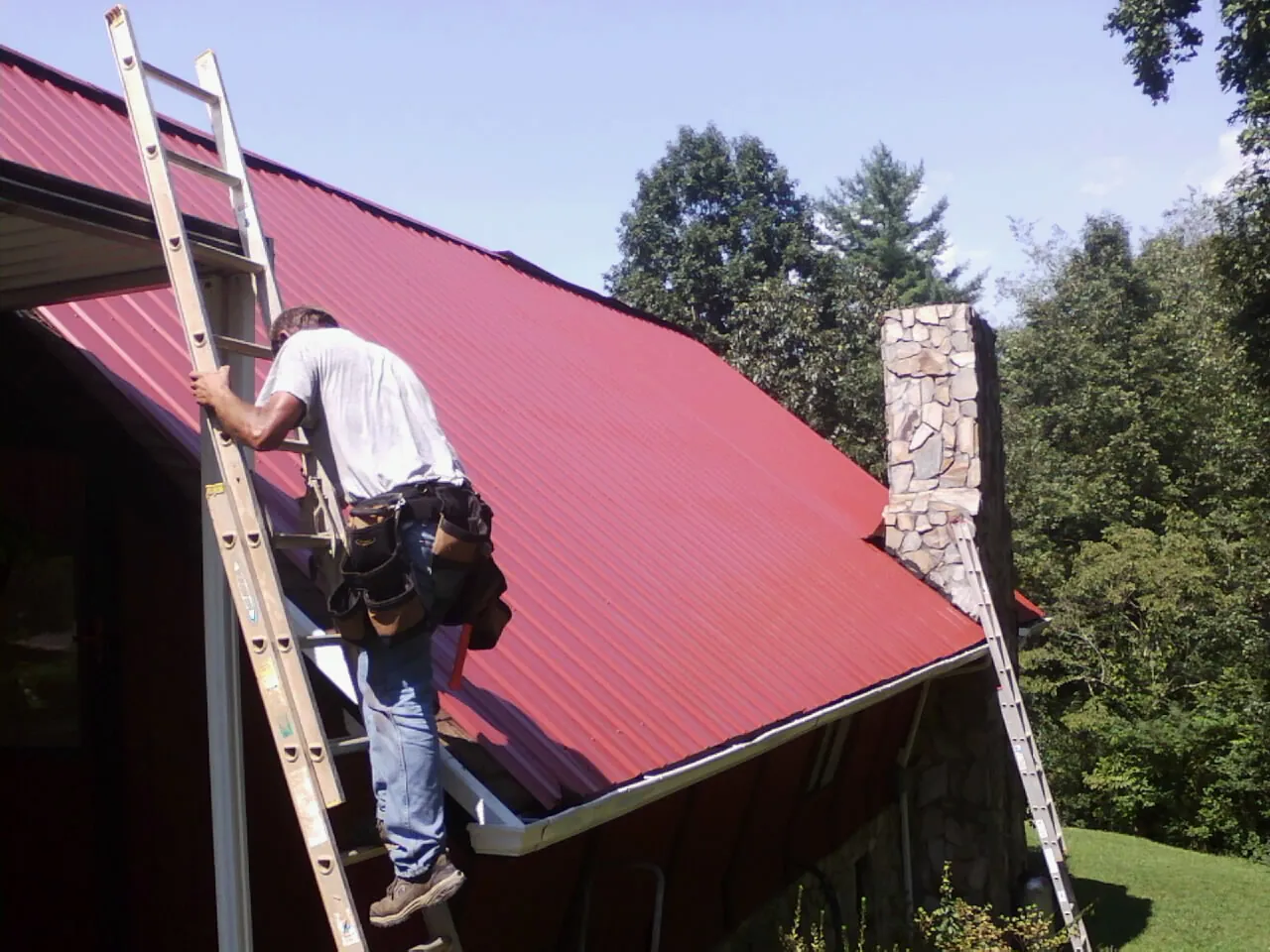 Roofer on a ladder beside a red metal roof and stone chimney — Soffit & Fascia Repair by Roofing Brothers in Ho Ho Kus