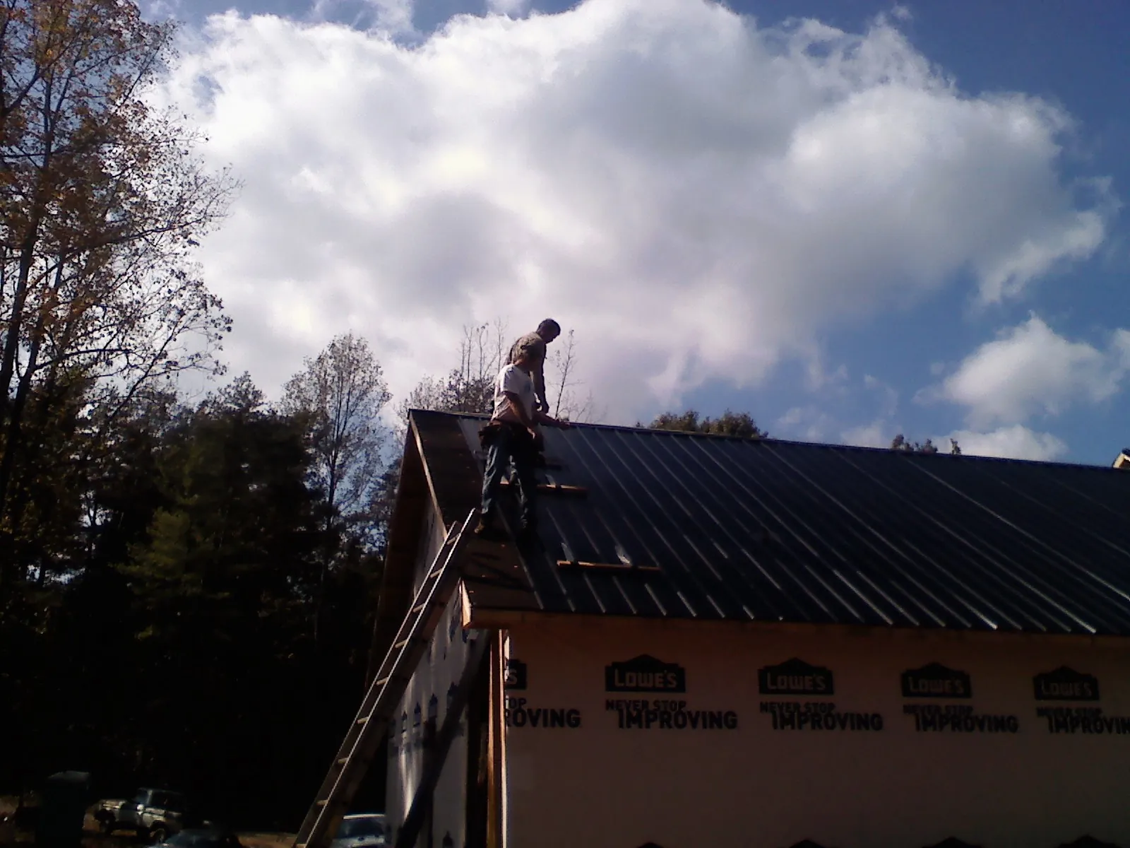 Roofer fastening dark metal panels high against a cloudy sky, from a Roof Inspection job in Ho Ho Kus